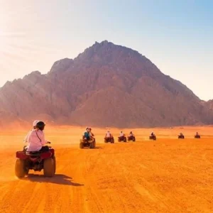 Gruppo di persone in quad che attraversano il deserto arancione di Marsa Alam al tramonto, con una grande montagna rocciosa sullo sfondo e il sole che illumina la scena