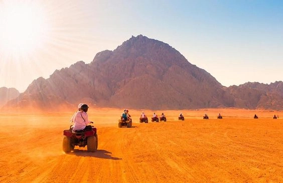 Gruppo di persone in quad che attraversano il deserto arancione di Marsa Alam al tramonto, con una grande montagna rocciosa sullo sfondo e il sole che illumina la scena