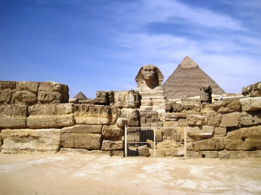 Il complesso delle Piramidi di Giza e la Sfinge sotto un cielo blu, con l'antica recinzione di pietra in primo piano e una stele.