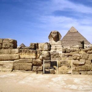 Il complesso delle Piramidi di Giza e la Sfinge sotto un cielo blu, con l'antica recinzione di pietra in primo piano e una stele.