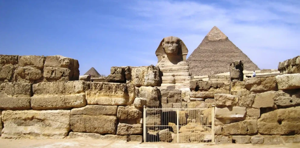Il complesso delle Piramidi di Giza e la Sfinge sotto un cielo blu, con l'antica recinzione di pietra in primo piano e una stele.