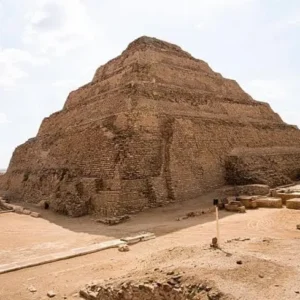 Una vista a livello del suolo della monumentale Piramide a Gradoni di Djoser (o Piramide di Saqqara), con la sua distinta struttura a sei livelli di pietra irregolare ocra, situata in un vasto deserto sabbioso pianeggiante. In primo piano, il terreno di sabbia compattata, resti di fondamenta antiche, pietre e un piccolo palo di marcatore. Lo sfondo mostra l'orizzonte piatto del deserto sotto un cielo luminoso con nuvole sparse e sfocate. La piramide è il punto focale, imponente e ben definita.