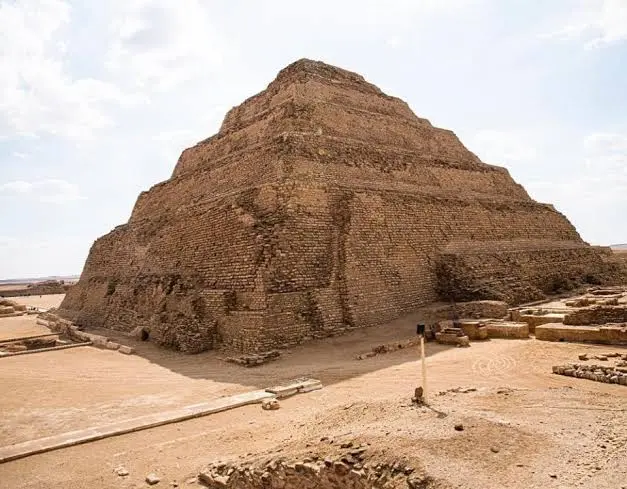 Una vista a livello del suolo della monumentale Piramide a Gradoni di Djoser (o Piramide di Saqqara), con la sua distinta struttura a sei livelli di pietra irregolare ocra, situata in un vasto deserto sabbioso pianeggiante. In primo piano, il terreno di sabbia compattata, resti di fondamenta antiche, pietre e un piccolo palo di marcatore. Lo sfondo mostra l'orizzonte piatto del deserto sotto un cielo luminoso con nuvole sparse e sfocate. La piramide è il punto focale, imponente e ben definita.