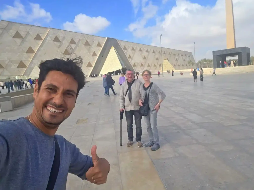 Un selfie di gruppo sorridente all'ingresso del nuovo Grand Egyptian Museum (GEM) con un turista che fa il segno "ok". Dietro di loro si vede l'architettura moderna del museo e l'obelisco all'aperto. Un momento felice all'inizio di una visita durante il tour del Cairo 5 giorni