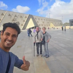 Un selfie di gruppo sorridente all'ingresso del nuovo Grand Egyptian Museum (GEM) con un turista che fa il segno "ok". Dietro di loro si vede l'architettura moderna del museo e l'obelisco all'aperto. Un momento felice all'inizio di una visita durante il tour del Cairo 5 giorni