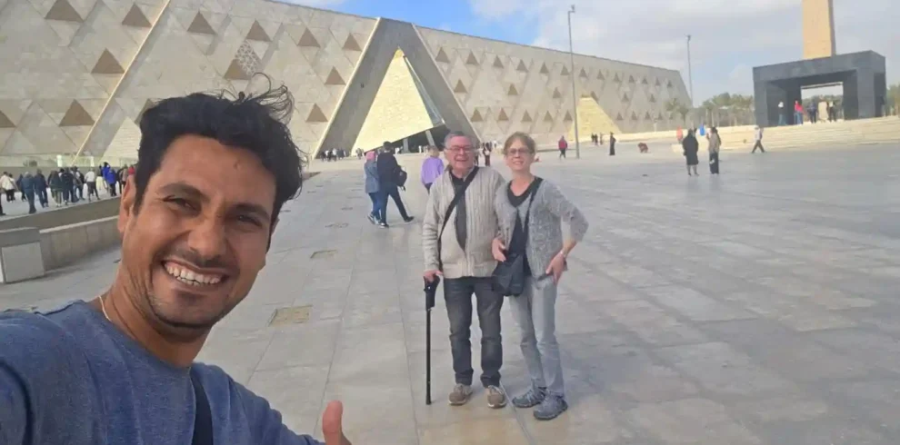 Un selfie di gruppo sorridente all'ingresso del nuovo Grand Egyptian Museum (GEM) con un turista che fa il segno "ok". Dietro di loro si vede l'architettura moderna del museo e l'obelisco all'aperto. Un momento felice all'inizio di una visita durante il tour del Cairo 5 giorni