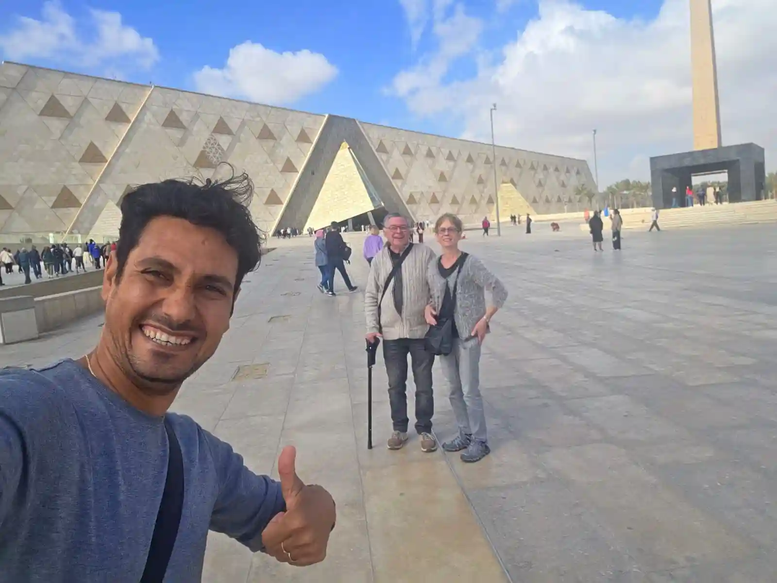 Un selfie di gruppo sorridente all'ingresso del nuovo Grand Egyptian Museum (GEM) con un turista che fa il segno "ok". Dietro di loro si vede l'architettura moderna del museo e l'obelisco all'aperto. Un momento felice all'inizio di una visita durante il tour del Cairo 5 giorni